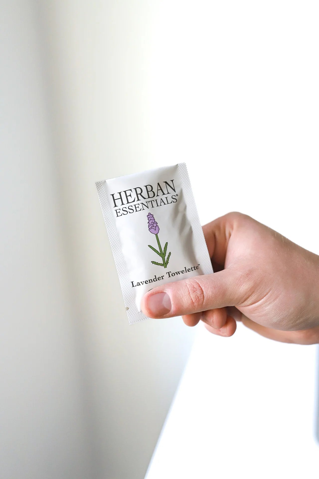 A hand holds a small packet labeled "Herban Essentials Essential Oil Towelettes," featuring a lavender sprig, against a plain light background—ideal for refreshing on the go.