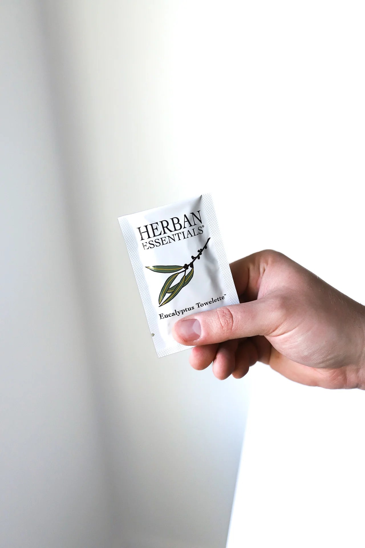 A hand holds a packet of Herban Essentials Essential Oil Towelettes, featuring a white design with the product name and an illustration of a eucalyptus branch on the front.