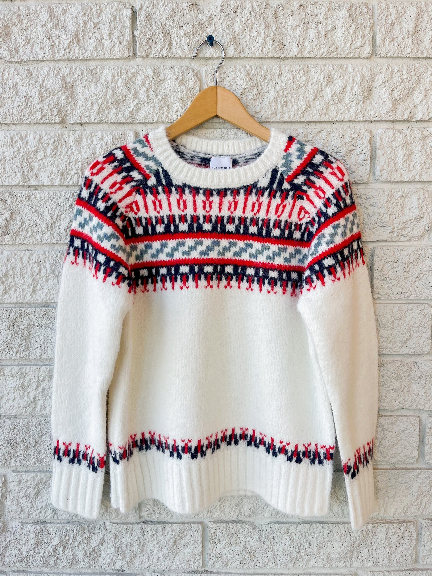 The Josie Sweater by Hunter Bell NYC is a white wool blend with red, navy, and black geometric fair isle patterns on the yoke, cuffs, and hem. Shown true to size on a wooden hanger against a white brick wall.