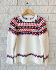 The Josie Sweater by Hunter Bell NYC is a white wool blend with red, navy, and black geometric fair isle patterns on the yoke, cuffs, and hem. Shown true to size on a wooden hanger against a white brick wall.