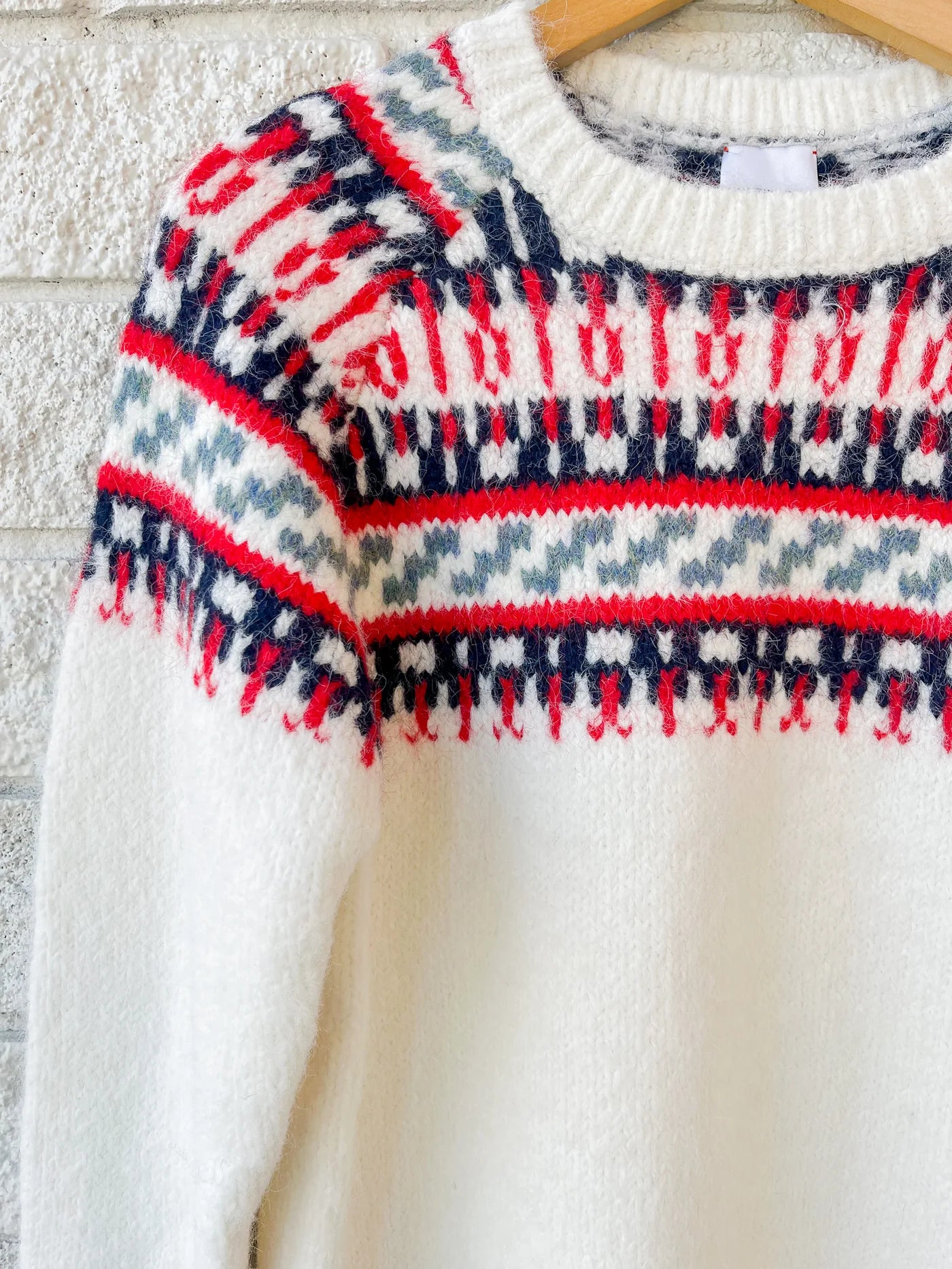 The Josie Sweater by Hunter Bell NYC, a cozy white wool-blend knit featuring red, navy, and gray Nordic patterns on the chest and shoulders, hangs on a wooden hanger against a white brick wall.
