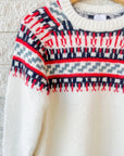 The Josie Sweater by Hunter Bell NYC, a cozy white wool-blend knit featuring red, navy, and gray Nordic patterns on the chest and shoulders, hangs on a wooden hanger against a white brick wall.