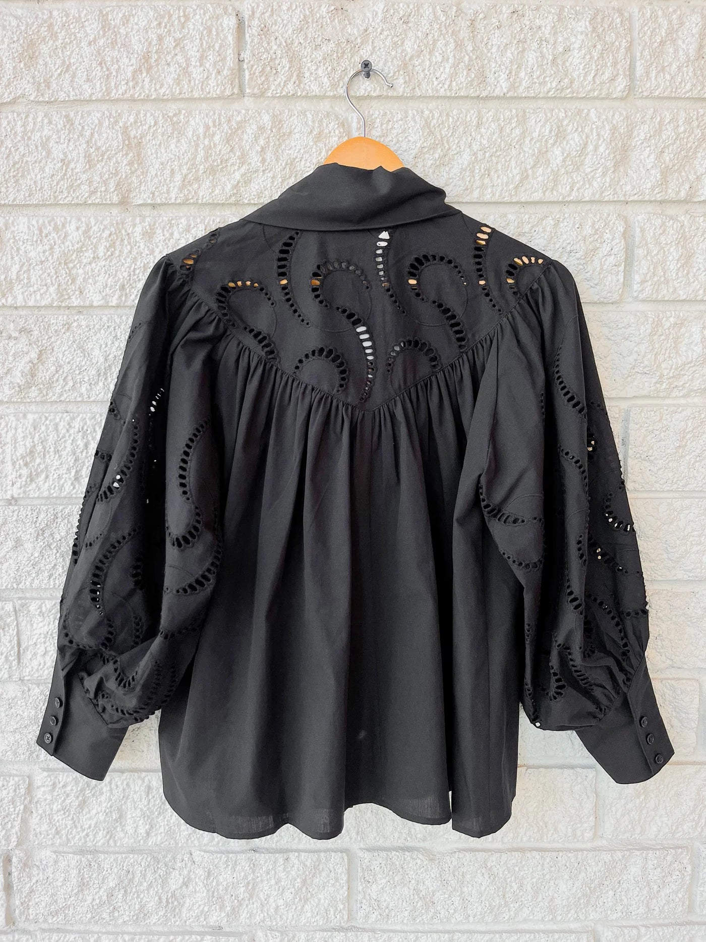 The Shay Blouse by Hunter Bell NYC hangs on a wooden hanger against a white brick wall, showcasing swirling shoulder embroidery and spacious sleeves. Gathered fabric ensures a relaxed, loose fit for effortless style.