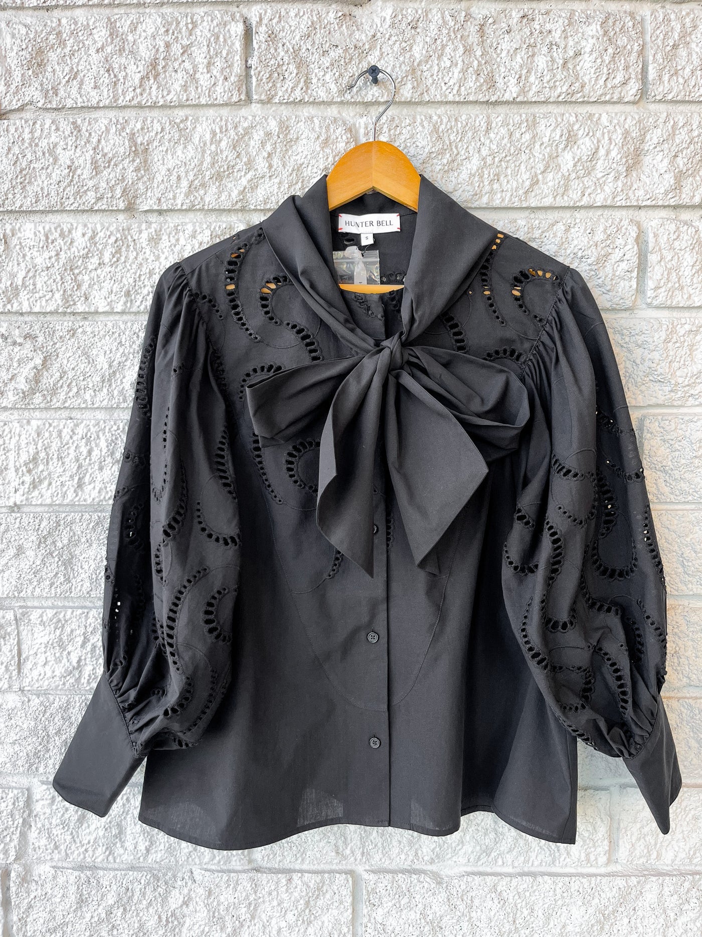 The Shay Blouse by Hunter Bell NYC, featuring black eyelet fabric, roomy sleeves, a button-down front, and a standout bow collar, is displayed on a wooden hanger against a white brick wall.
