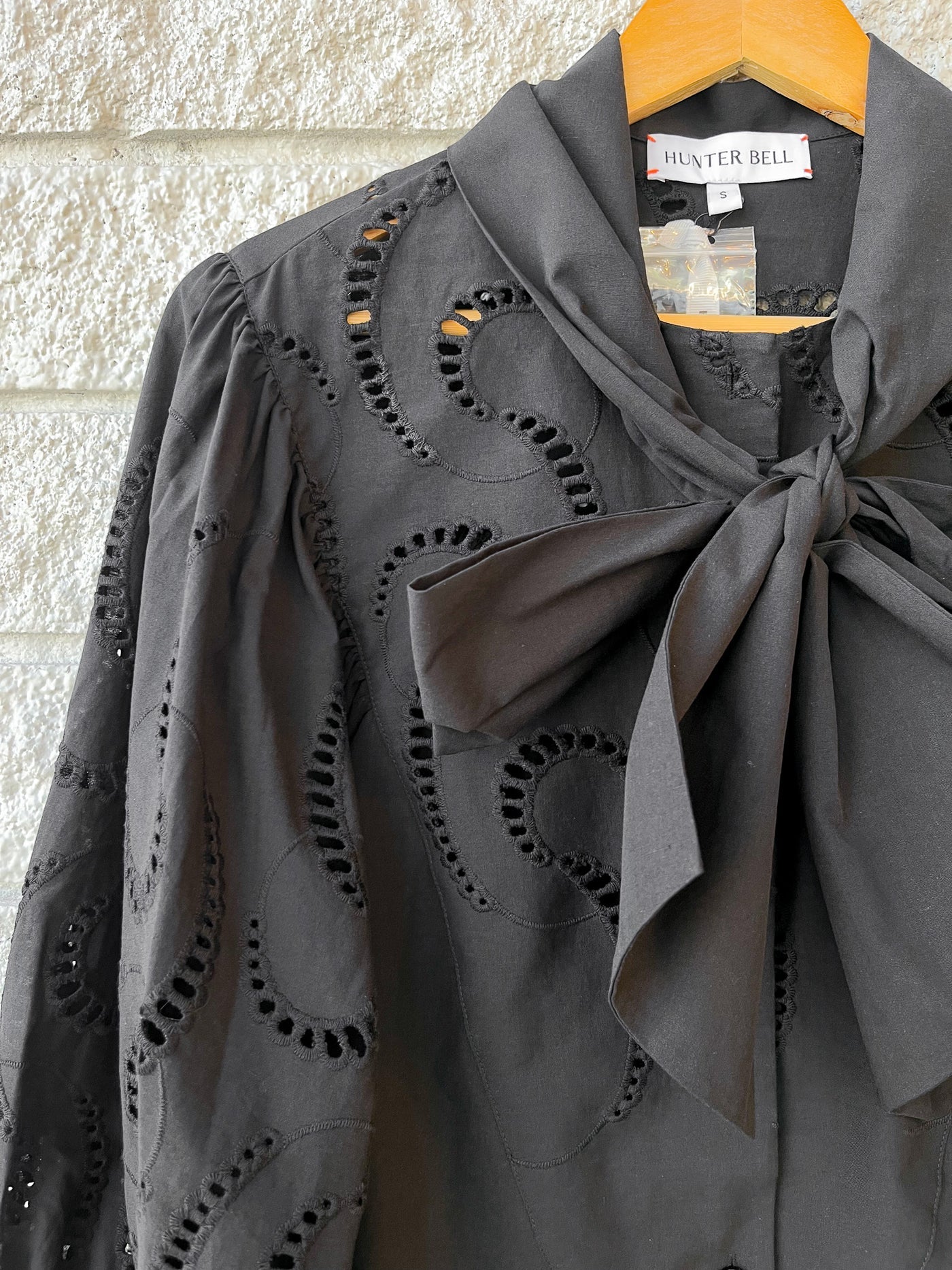 Close-up of the Shay Blouse by Hunter Bell NYC—a black eyelet top with spacious sleeves and a dramatic bow collar, hanging on a wooden hanger against a textured light stone wall.