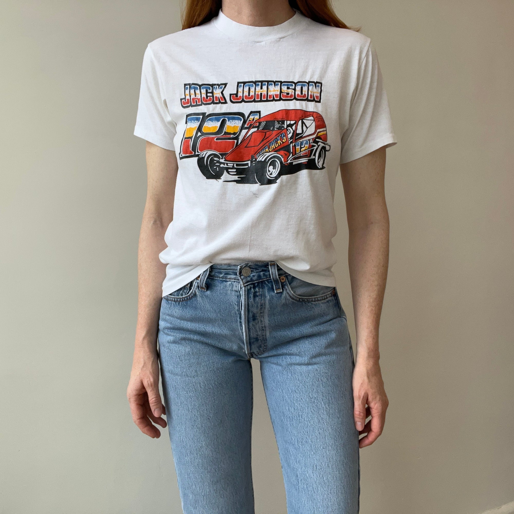 A person wearing the 80s Jack Johnson Drag Racing Tee from Red Vintage Co., featuring a red racing car graphic and "Jack Johnson" text, stands against a plain background. The single stitch detailing gives their outfit a classic touch, paired perfectly with light blue jeans.