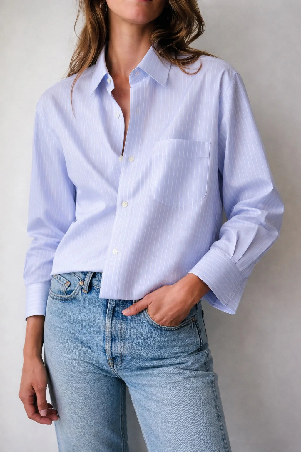 A woman wears Cissa's The Shorty Shirt in light blue Egyptian cotton with thin Italian stripes, styled slightly oversized, paired with light blue jeans. She stands against a plain background with one hand in her pocket.