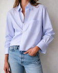 A woman wears Cissa's The Shorty Shirt in light blue Egyptian cotton with thin Italian stripes, styled slightly oversized, paired with light blue jeans. She stands against a plain background with one hand in her pocket.