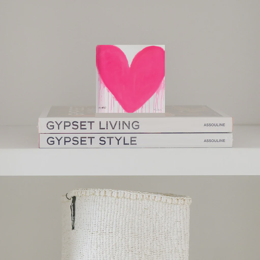 A minimalist shelf displays two stacked books, titled "Gypset Living" and "Gypset Style" by Assouline. On top of the books is an 8x8 canvas block featuring a bright pink, dripping heart by Kerri Rosenthal, called "In My Heart Drippy Heart Block". Below the shelf, a woven white basket is partially visible.