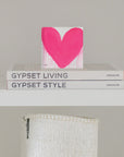 A minimalist shelf displays two stacked books, titled "Gypset Living" and "Gypset Style" by Assouline. On top of the books is an 8x8 canvas block featuring a bright pink, dripping heart by Kerri Rosenthal, called "In My Heart Drippy Heart Block". Below the shelf, a woven white basket is partially visible.