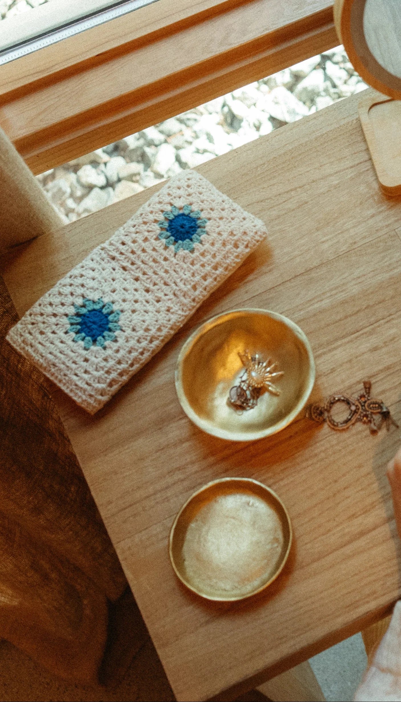 An Indaba 1-1234 Forged Brass Bowl showcases jewelry, like a sun-shaped ring, on a wooden table beside a white crocheted pouch with blue flowers, near a sunny window—stylish and elegant for desk organization.