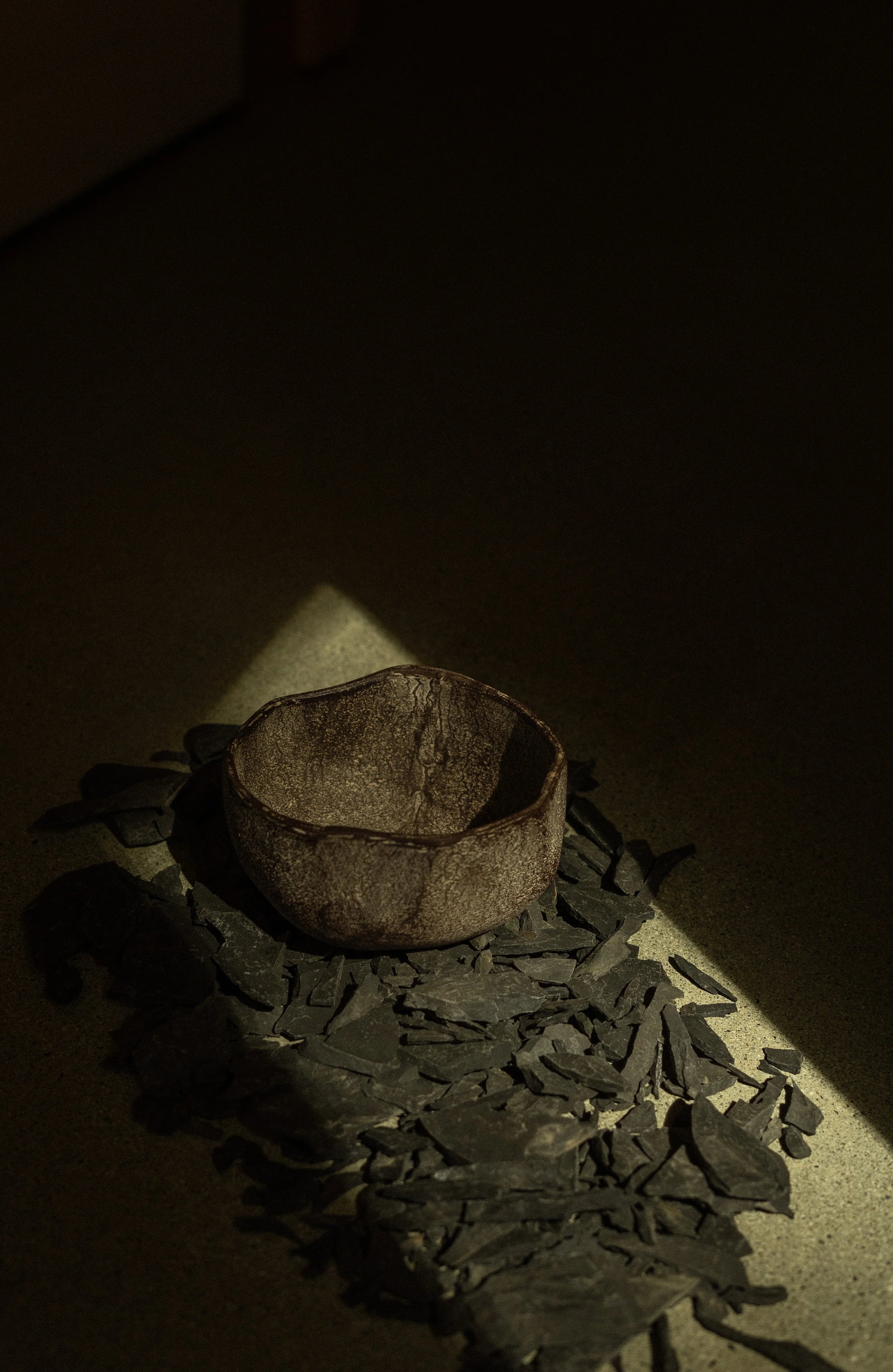 The Rustica Bowl S by Indaba—a small, handcrafted aluminum bowl made in India using sand casting—rests on dark slate shards, highlighted by a beam of light against a shadowy background.