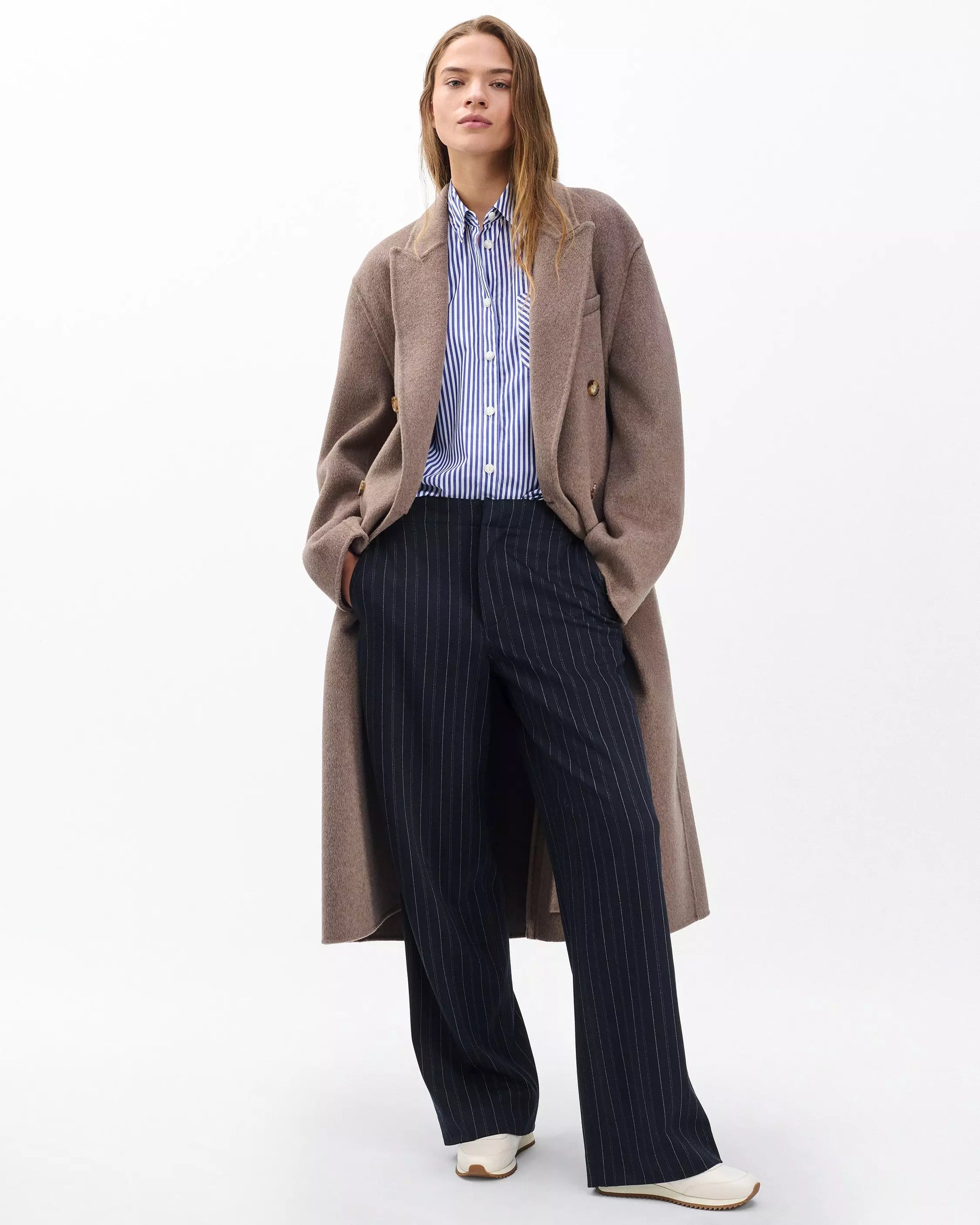 A woman wears Rag & Bone’s irina ponte tailored pant in navy, paired with white shoes, a blue-and-white striped button-up shirt, and a long taupe coat, standing against a plain white background.