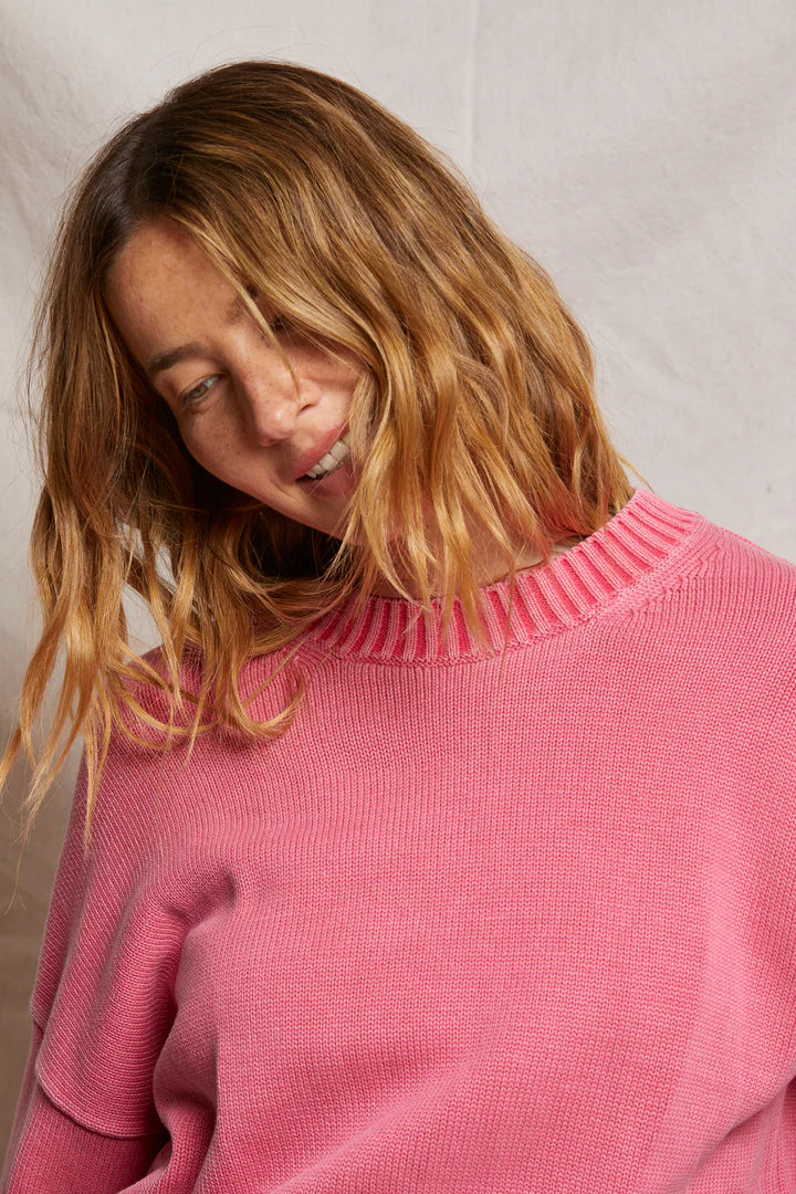 A person with wavy, shoulder-length hair smiles, head tilted slightly while wearing the Janet Sweater by Perfectwhitetee—a pink cotton knit with a textured collar and wardrobe staple. The backdrop is a simple, light-colored fabric.