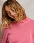 A person with wavy, shoulder-length hair smiles, head tilted slightly while wearing the Janet Sweater by Perfectwhitetee—a pink cotton knit with a textured collar and wardrobe staple. The backdrop is a simple, light-colored fabric.