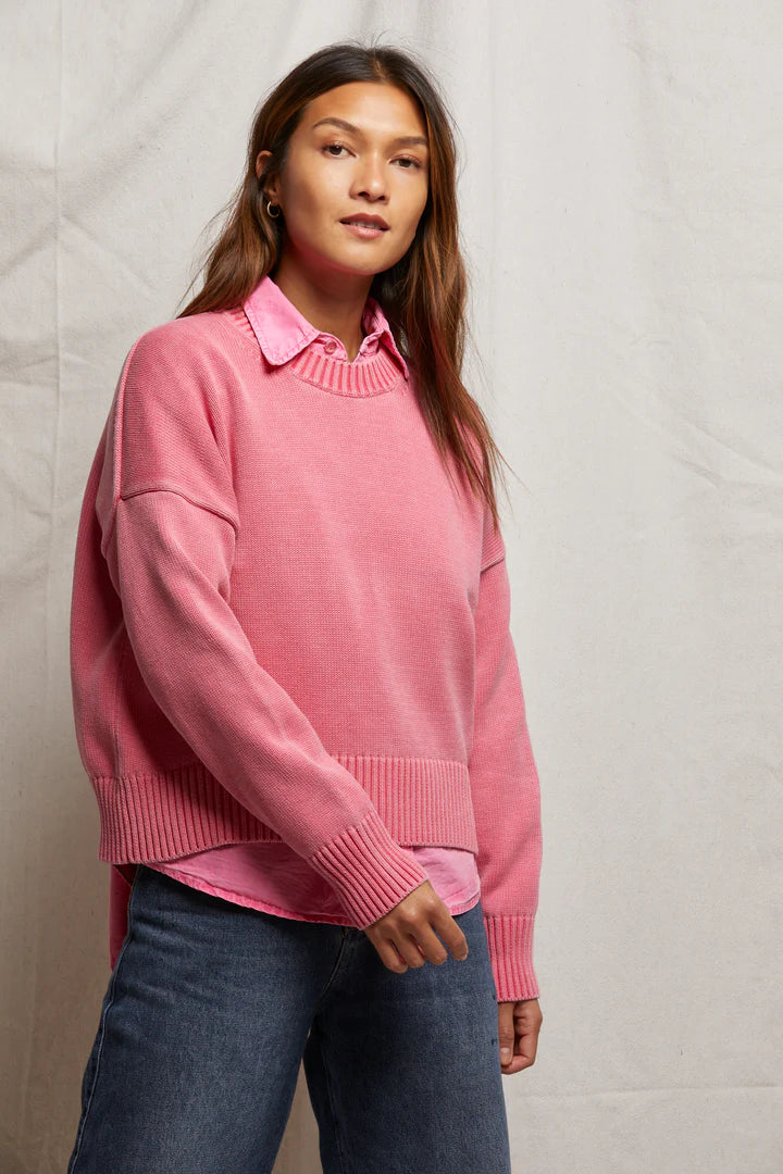 A woman in the boxy Janet Sweater by Perfectwhitetee, layered over a collared shirt and jeans, stands against a neutral fabric backdrop while gazing slightly aside. This sweater adds effortless style to her look.