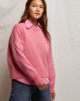 A woman in the boxy Janet Sweater by Perfectwhitetee, layered over a collared shirt and jeans, stands against a neutral fabric backdrop while gazing slightly aside. This sweater adds effortless style to her look.