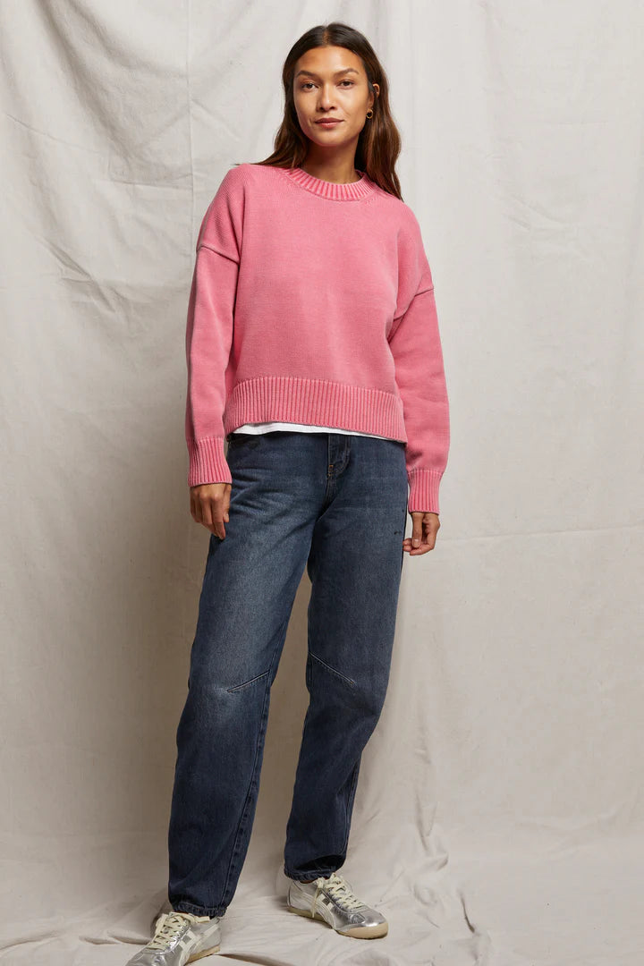 A person stands against a plain backdrop wearing the Janet Sweater by Perfectwhitetee, a boxy pink staple layered over a white shirt. Paired with blue jeans and gray sneakers, their long hair and relaxed posture showcase effortless style.