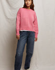 A person stands against a plain backdrop wearing the Janet Sweater by Perfectwhitetee, a boxy pink staple layered over a white shirt. Paired with blue jeans and gray sneakers, their long hair and relaxed posture showcase effortless style.