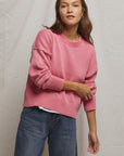 A person with long, dark hair wearing the Janet Sweater by Perfectwhitetee, in a pink boxy style, pairs it with blue jeans against a neutral backdrop. They gently touch their arm, highlighting the sweater's comfort as a staple knit.