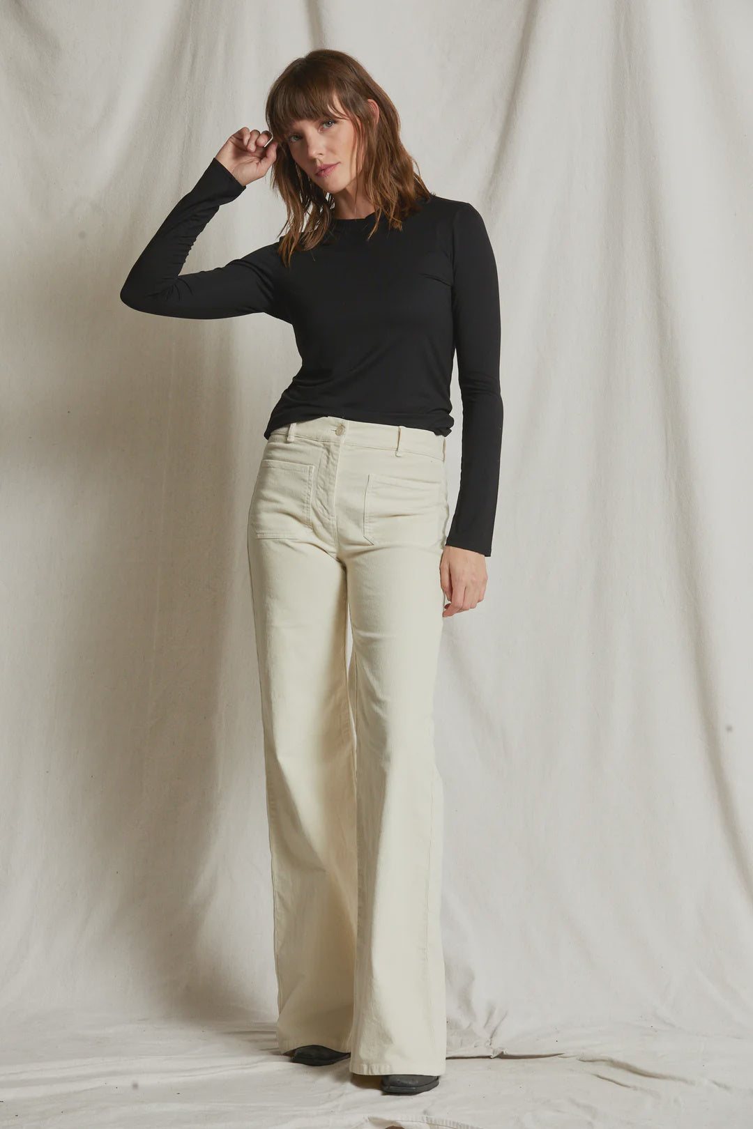 A person with shoulder-length hair stands against a plain, light-colored backdrop, exuding smooth '90s vibes in a black Dionne LS Baby Tee by Perfectwhitetee and high-waisted, wide-leg cream-colored pants. One hand is resting on their head while the other hangs by their side. Perfect fall and winter style.