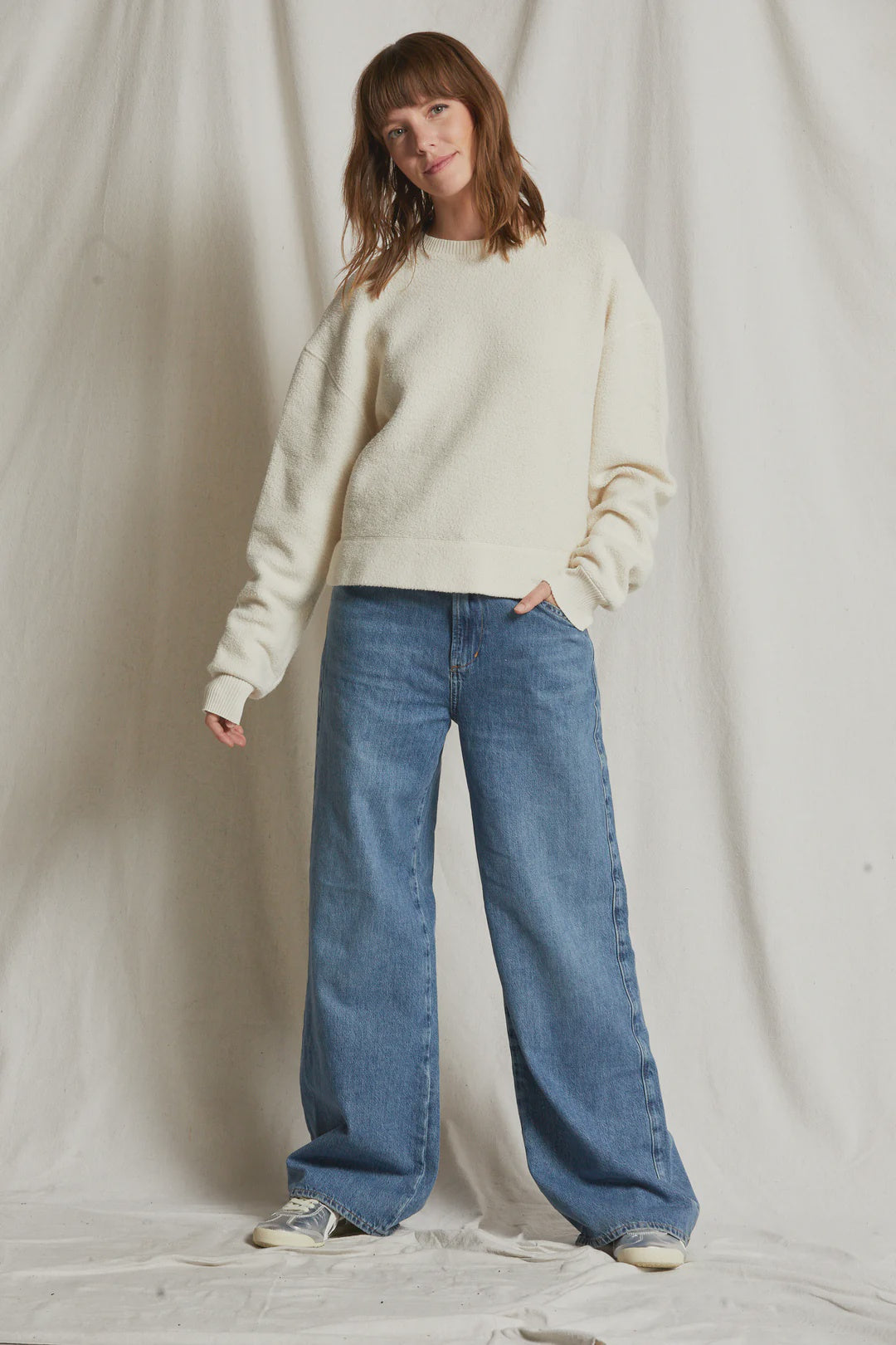 A woman stands against a plain, light-colored backdrop. She is wearing a vintage look: the Perfectwhitetee Mayer LS reverse fleece, a cream-colored, relaxed boxy sweatshirt with long sleeves, paired with loose-fitting blue jeans and white sneakers. She has a relaxed posture with one hand by her side and the other slightly raised.