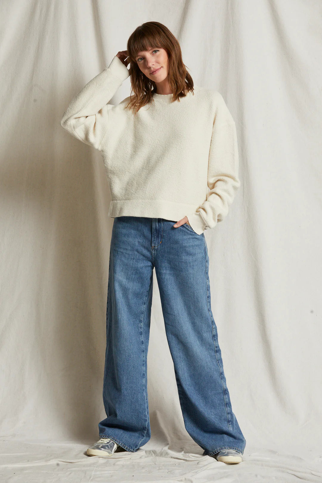 A person stands in front of a neutral fabric background, wearing the Mayer LS reverse fleece by Perfectwhitetee, characterized by its white luxe fluff material and boxy relaxed fit, paired with loose-fitting blue jeans. Their right hand rests at their side while their left hand touches their hair. They are also wearing sneakers and smiling slightly.