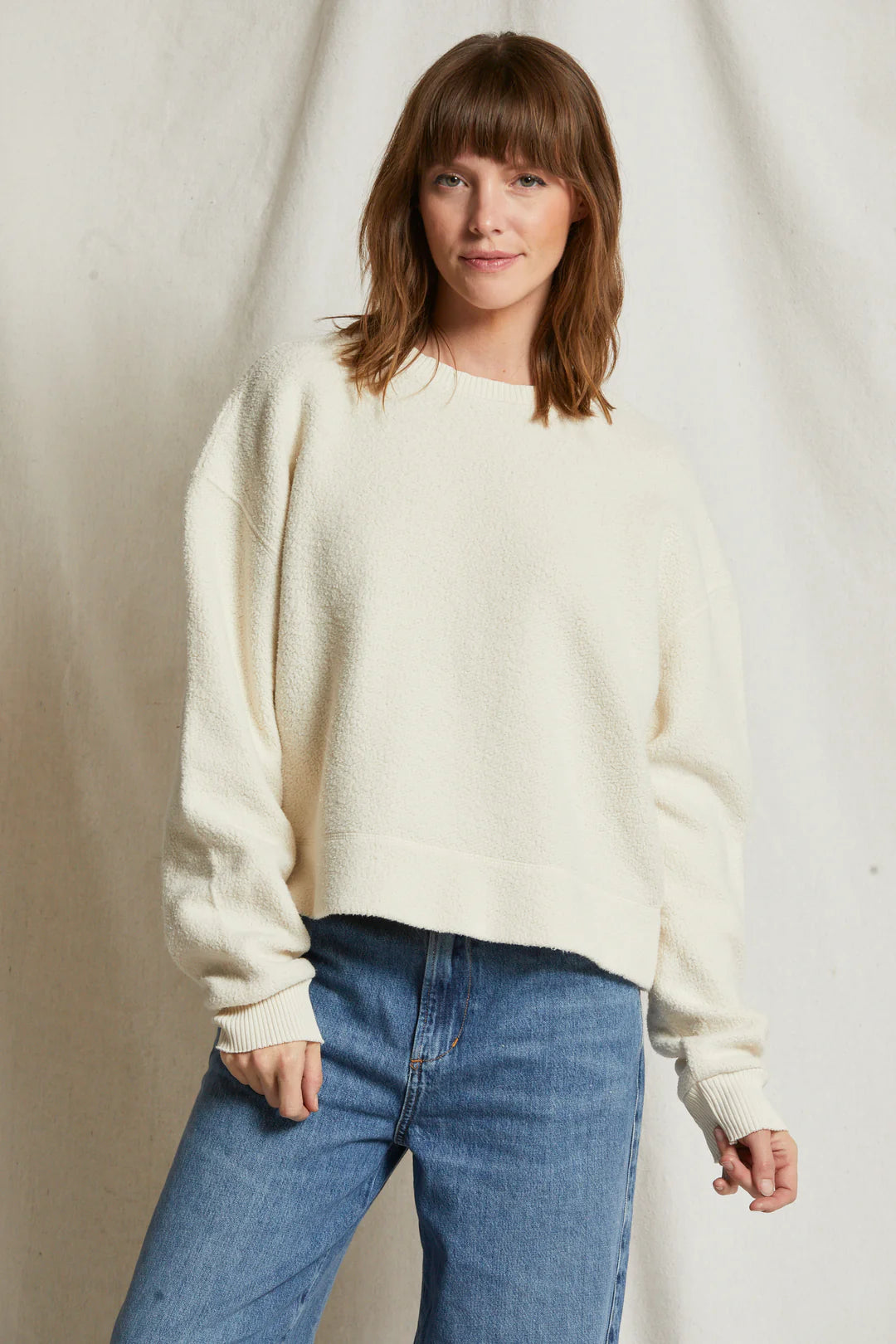 A person with shoulder-length brown hair stands against a plain background, wearing the Mayer LS reverse fleece sweater from Perfectwhitetee and blue jeans. They are posing casually with one hand resting on their hip and a slight smile on their face, giving the scene a vintage feel.