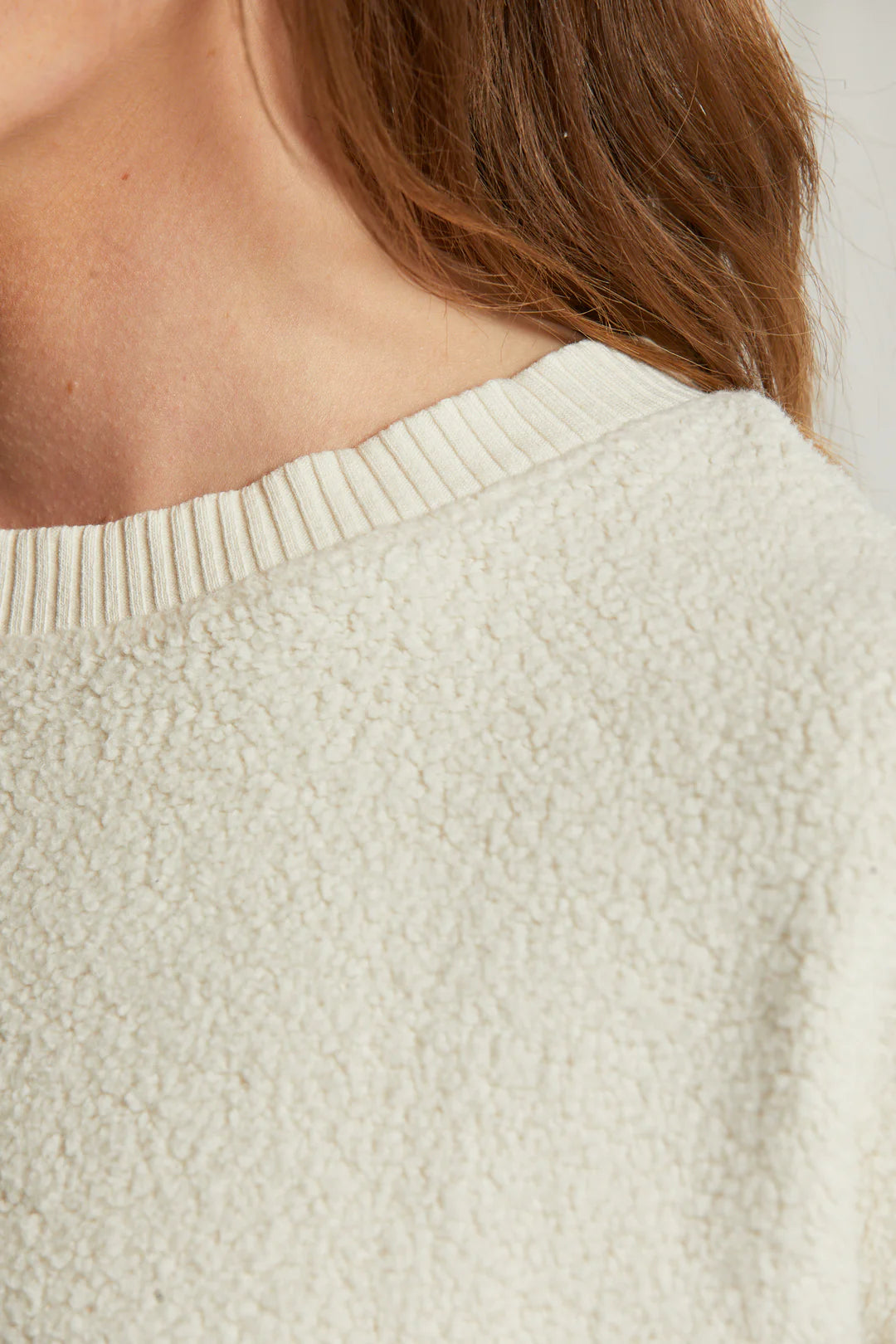 Close-up of a person&#39;s shoulder and neck wearing the Mayer LS reverse fleece by Perfectwhitetee. This cream-colored, textured sweater offers a cozy and vintage feel. The fabric appears soft with a knit finish around the neckline, complemented by the person&#39;s long, brown hair partially resting over it.