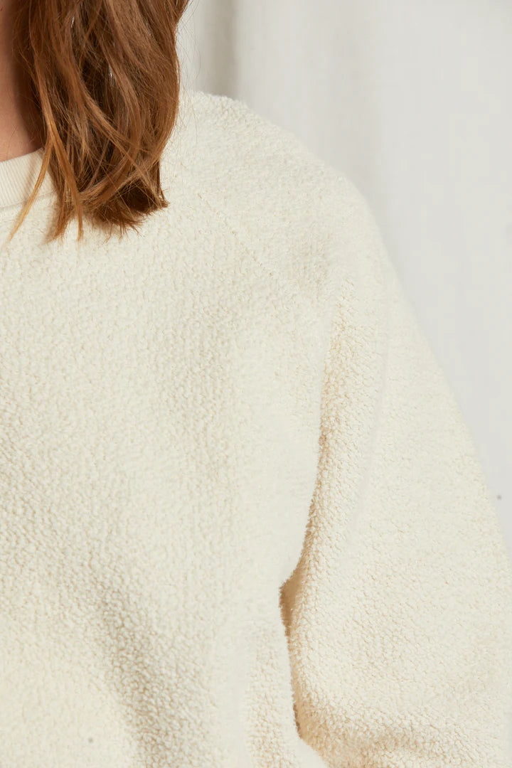 A close-up of someone wearing the Perfectwhitetee Ziggy Reverse LS Sweatshirt in off-white cotton fleece, with only their shoulder, arm, and brown hair visible against a plain background.