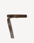 The Bowe Belt by Janessa Leone is crafted from Earth Brown regenerative leather, featuring a custom-designed rectangular brass buckle. It lays against a plain light background with multiple holes for adjustable sizing, and is made locally in Los Angeles.