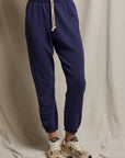 A person wearing Perfectwhitetee's French Terry Easy Sweatpants in navy blue, made from buttery soft cotton, pairs them with beige sneakers while standing on a draped fabric surface. These sweatpants feature a drawstring waist and are set against a backdrop of neutral-colored fabric curtains.