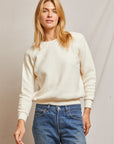 A person with long blonde hair, wearing a cozy Ziggy Reverse Fleece LS Crew Sweatshirt by Perfectwhitetee and blue jeans, stands in front of a plain background. They have a relaxed and confident expression, with one hand slightly raised.