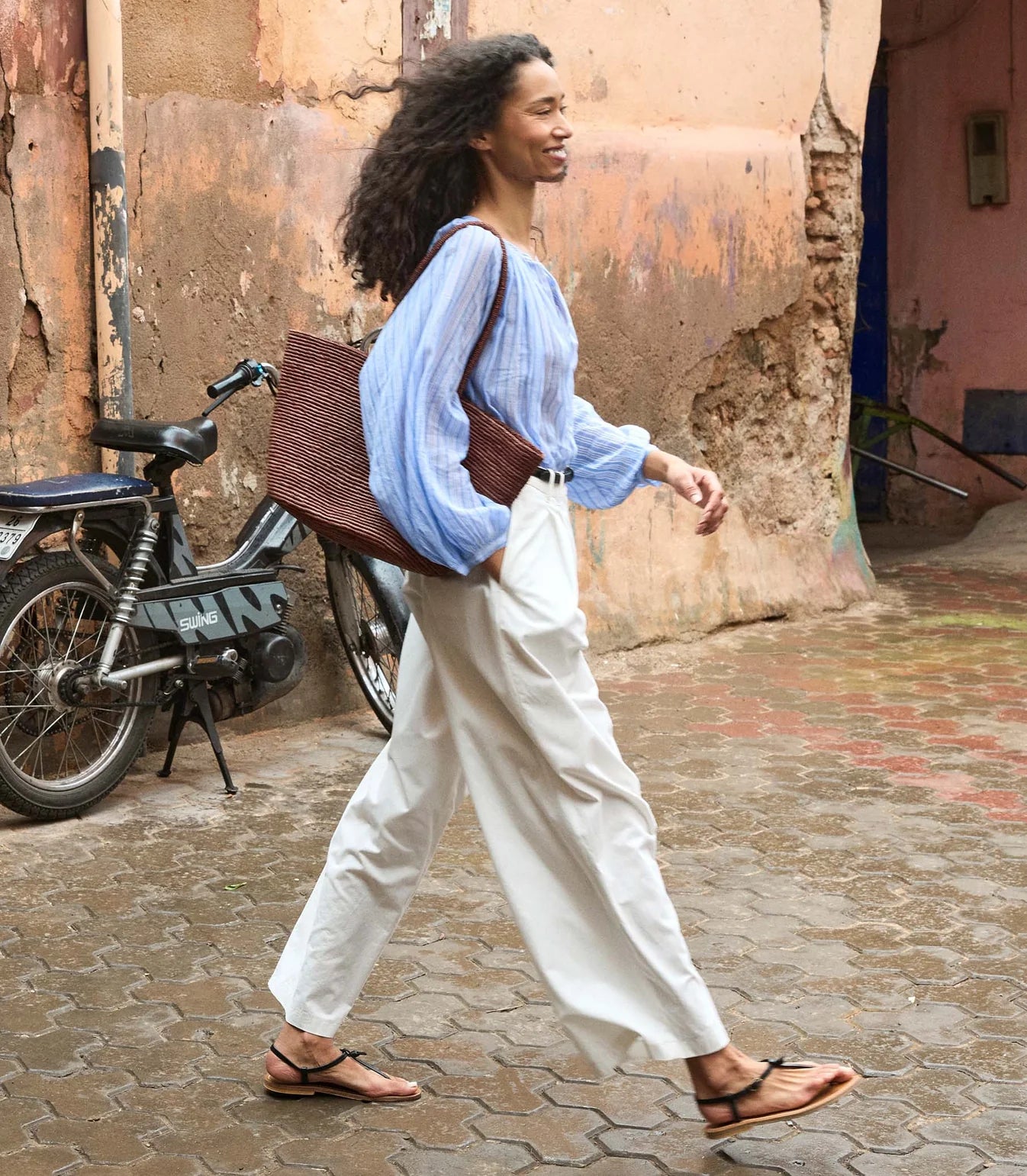 A woman with curly hair wearing the DOEN Jonie Top, white pants, and sandals walks past a weathered wall and parked motorcycle, carrying a large brown woven bag. The tiled ground is wet.