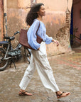 A woman with curly hair wearing the DOEN Jonie Top, white pants, and sandals walks past a weathered wall and parked motorcycle, carrying a large brown woven bag. The tiled ground is wet.