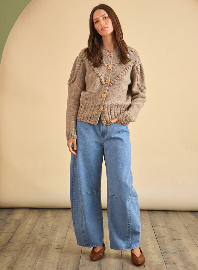 A woman with long brown hair stands indoors, facing forward in a neutral pose, wearing the Bobble Sweater by Hunter Bell NYC, wide-leg blue jeans, and brown shoes against a beige and green background.