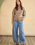 A woman with long brown hair stands indoors, facing forward in a neutral pose, wearing the Bobble Sweater by Hunter Bell NYC, wide-leg blue jeans, and brown shoes against a beige and green background.