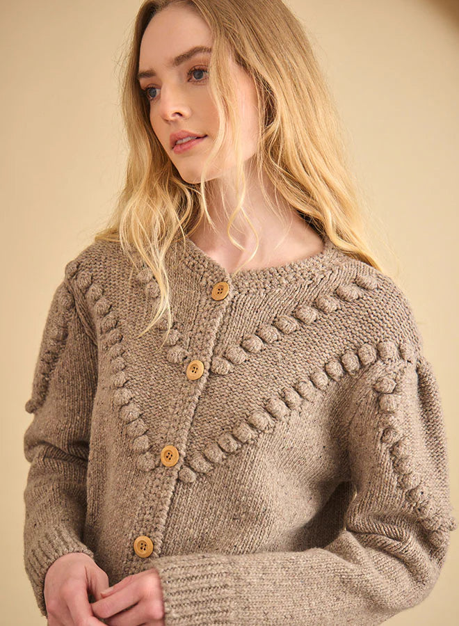 A woman with long blonde hair wears the Hunter Bell NYC Bobble Sweater, a textured light brown knit cardigan with wooden buttons and decorative patterns, standing against a beige background.