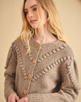 A woman with long blonde hair wears the Hunter Bell NYC Bobble Sweater, a textured light brown knit cardigan with wooden buttons and decorative patterns, standing against a beige background.
