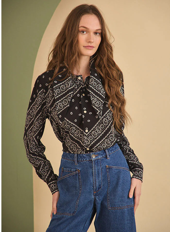 A woman with long wavy brown hair wears the Hunter Bell NYC Frances Blouse, a black patterned long-sleeve top, and high-waisted blue jeans, standing in front of a beige and green background.