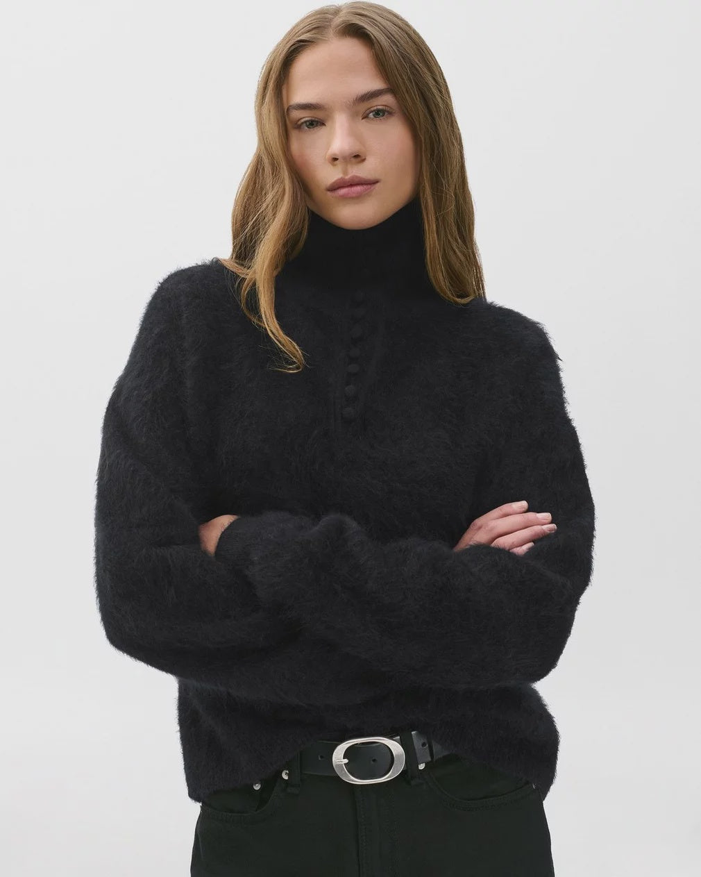 A woman with long, light brown hair stands against a plain background, wearing the oversized-fit jada brushed cashmere sweater by Rag & Bone and black belted pants. Her arms are crossed and her expression is neutral.