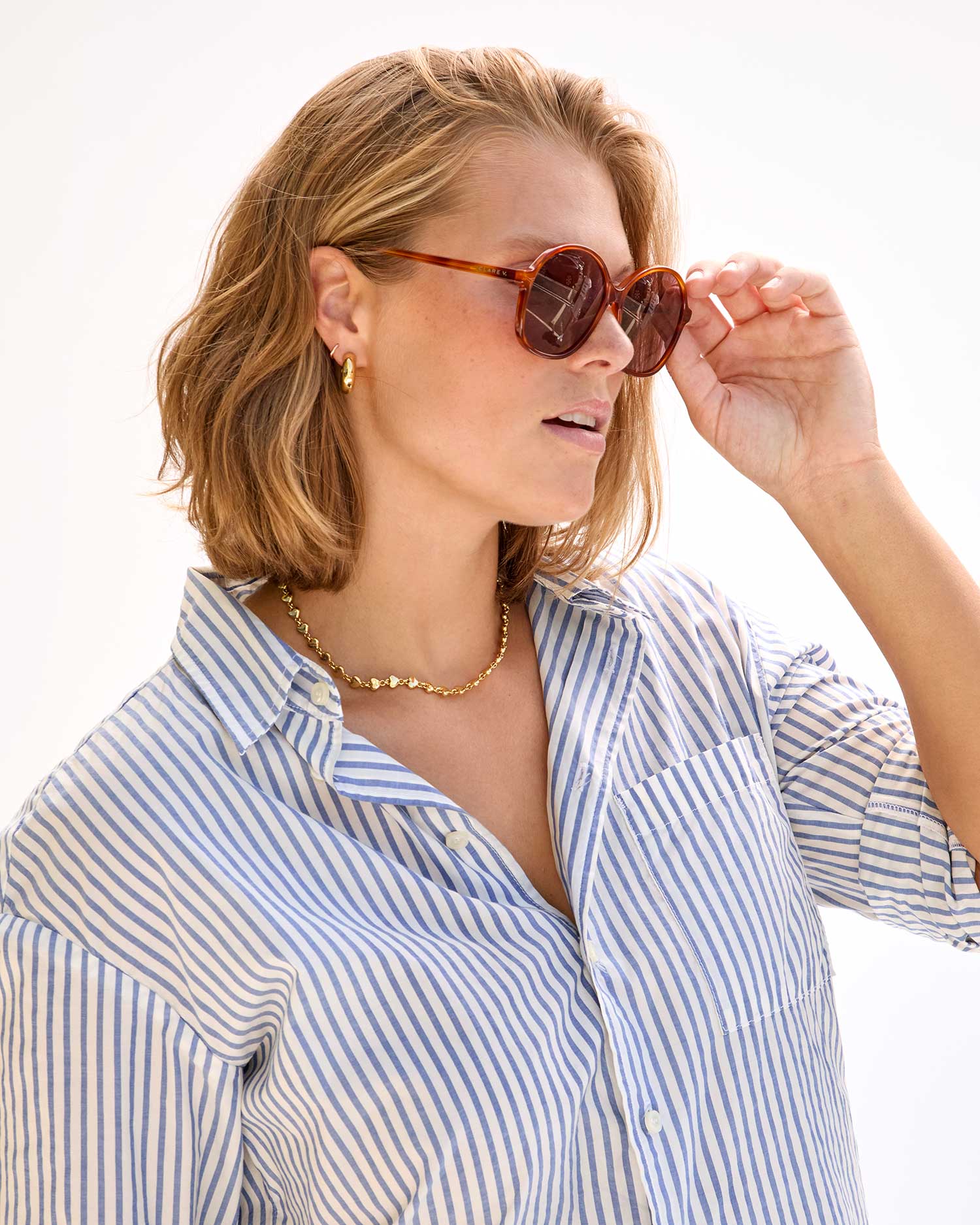 A person with shoulder-length blonde hair is wearing round Clare Vivier Jane Sunglasses in brown, which feature UVA/UVB protection. They are also dressed in a blue-and-white striped button-up shirt, accessorized with a gold necklace and gold hoop earrings. They are turned to the side, holding the sunglasses with one hand.