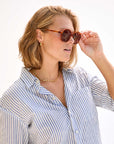 A person with shoulder-length blonde hair is wearing round Clare Vivier Jane Sunglasses in brown, which feature UVA/UVB protection. They are also dressed in a blue-and-white striped button-up shirt, accessorized with a gold necklace and gold hoop earrings. They are turned to the side, holding the sunglasses with one hand.