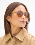 A woman with long brown hair wears amber-tinted Jane Sunglasses by Clare Vivier and a beige button-up shirt. She has a pair of small hoop earrings and looks to her left with a neutral expression. The background is plain and light-colored.