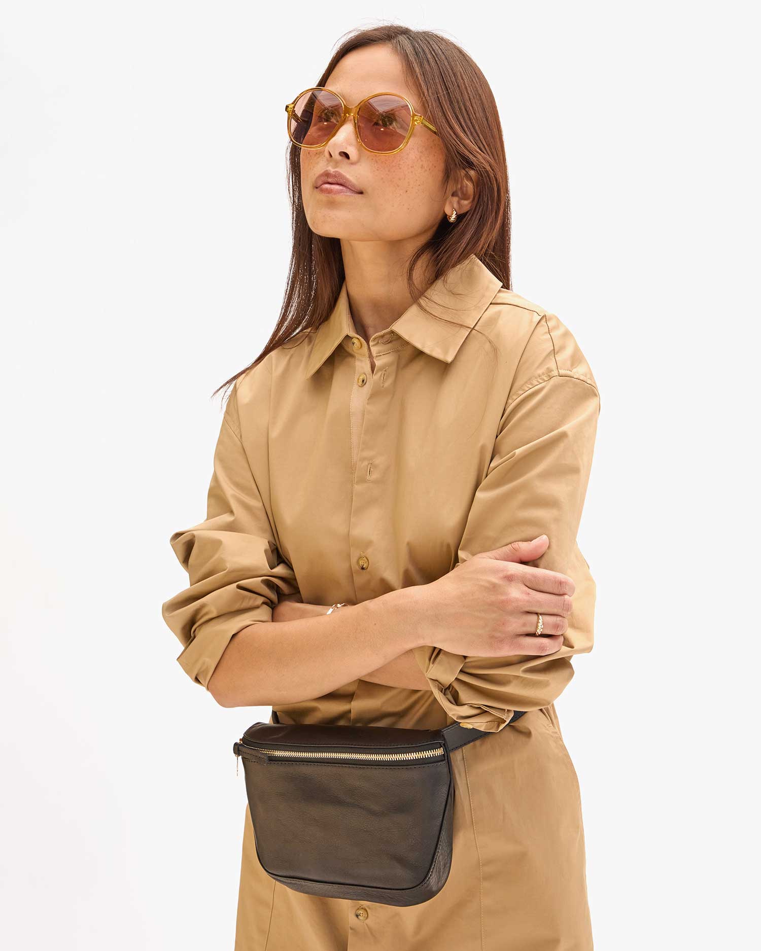 A person wearing a beige trench coat and Clare Vivier's Jane Sunglasses with UVA/UVB protection stands confidently against a plain background. They have their arms crossed and are sporting a black cross-body bag made from recycled materials.