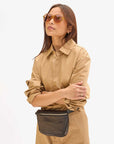 A woman in a tan button-up dress, yellow-tinted sunglasses, and the Clare Vivier Fanny Pack Fall 25—made from natural grain leather—stands with arms crossed against a plain white background.