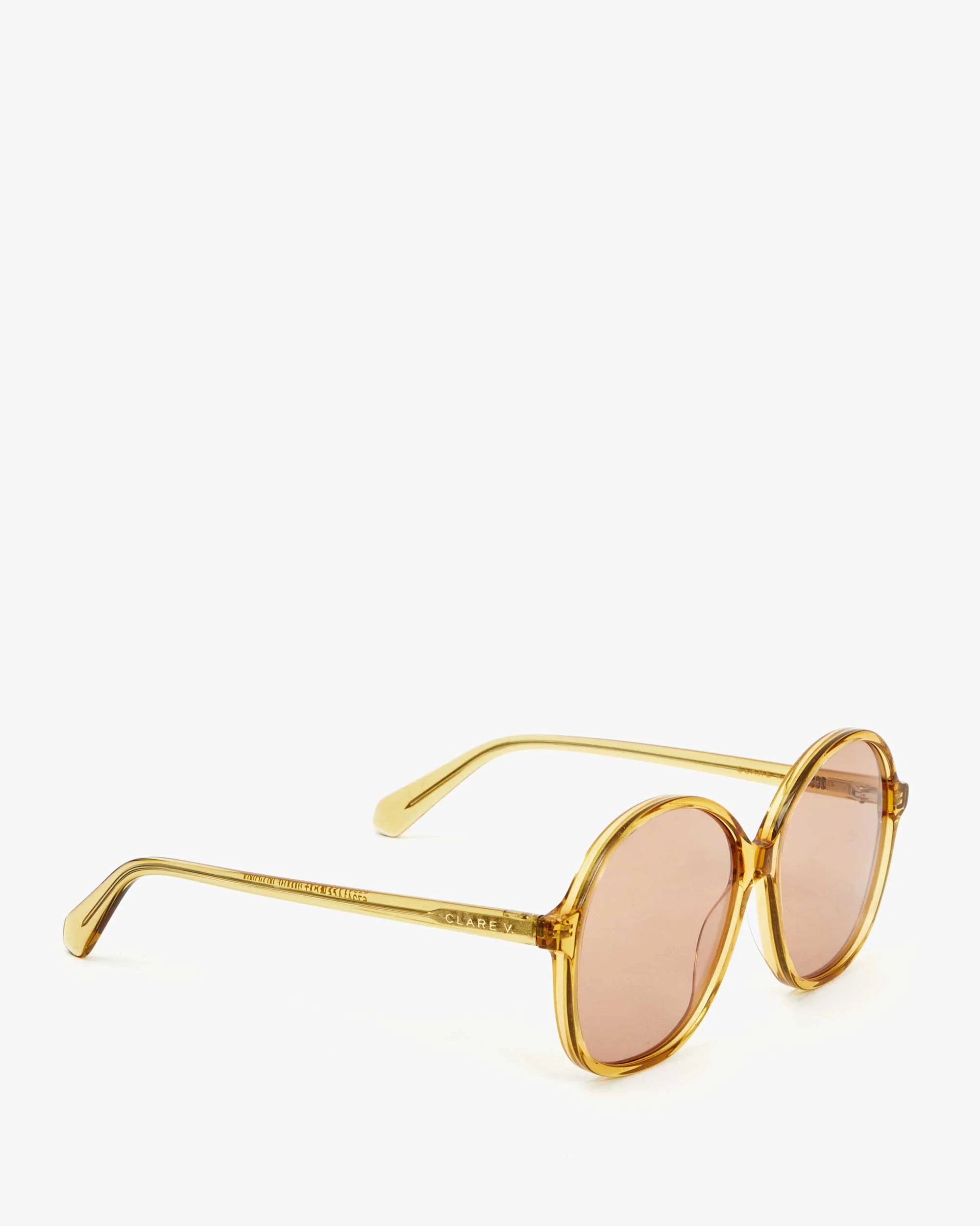 The Jane Sunglasses by Clare Vivier are a pair of vintage-style sunglasses with round, pink-tinted lenses and a golden frame. Crafted from recycled materials, the temples are engraved with text. Featuring UVA/UVB protection, the sunglasses are positioned with the lenses facing to the right on a white background.