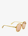 The Jane Sunglasses by Clare Vivier are a pair of vintage-style sunglasses with round, pink-tinted lenses and a golden frame. Crafted from recycled materials, the temples are engraved with text. Featuring UVA/UVB protection, the sunglasses are positioned with the lenses facing to the right on a white background.