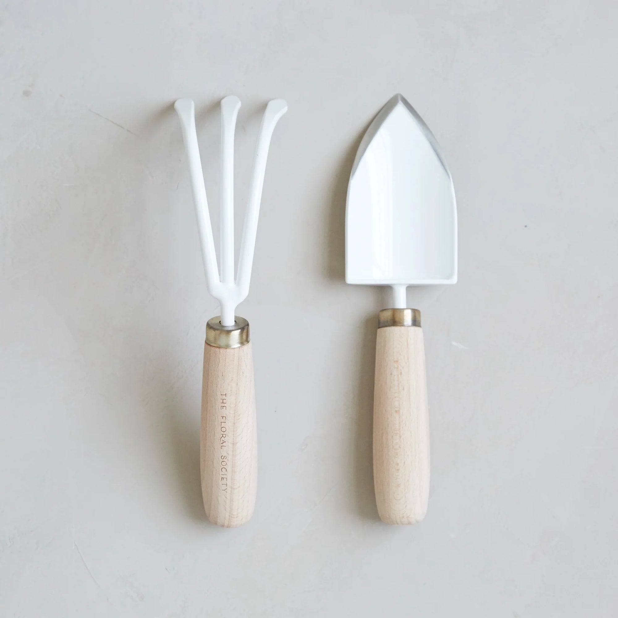 Displayed on a light surface is a compact Garden Tool Set by The Floral Society, showcasing a white garden trowel and hand rake. Both tools are meticulously crafted from Japanese stainless steel, complemented by light wooden handles adorned with metal collars, emphasizing their attention to detail and quality craftsmanship.