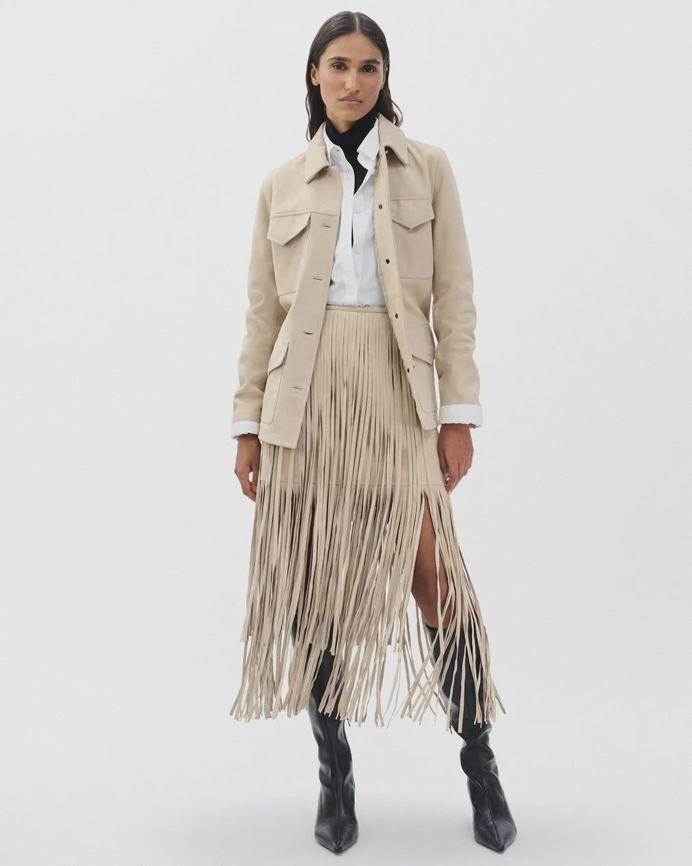 A woman stands against a plain background wearing a beige suede jacket, a white button-up layered over a black turtleneck, the Rag & Bone jasmine fringe skirt, and black heeled boots.